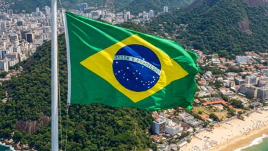 A photo of a large Brazilian flag waving in the wind in front of a scenic landscape.