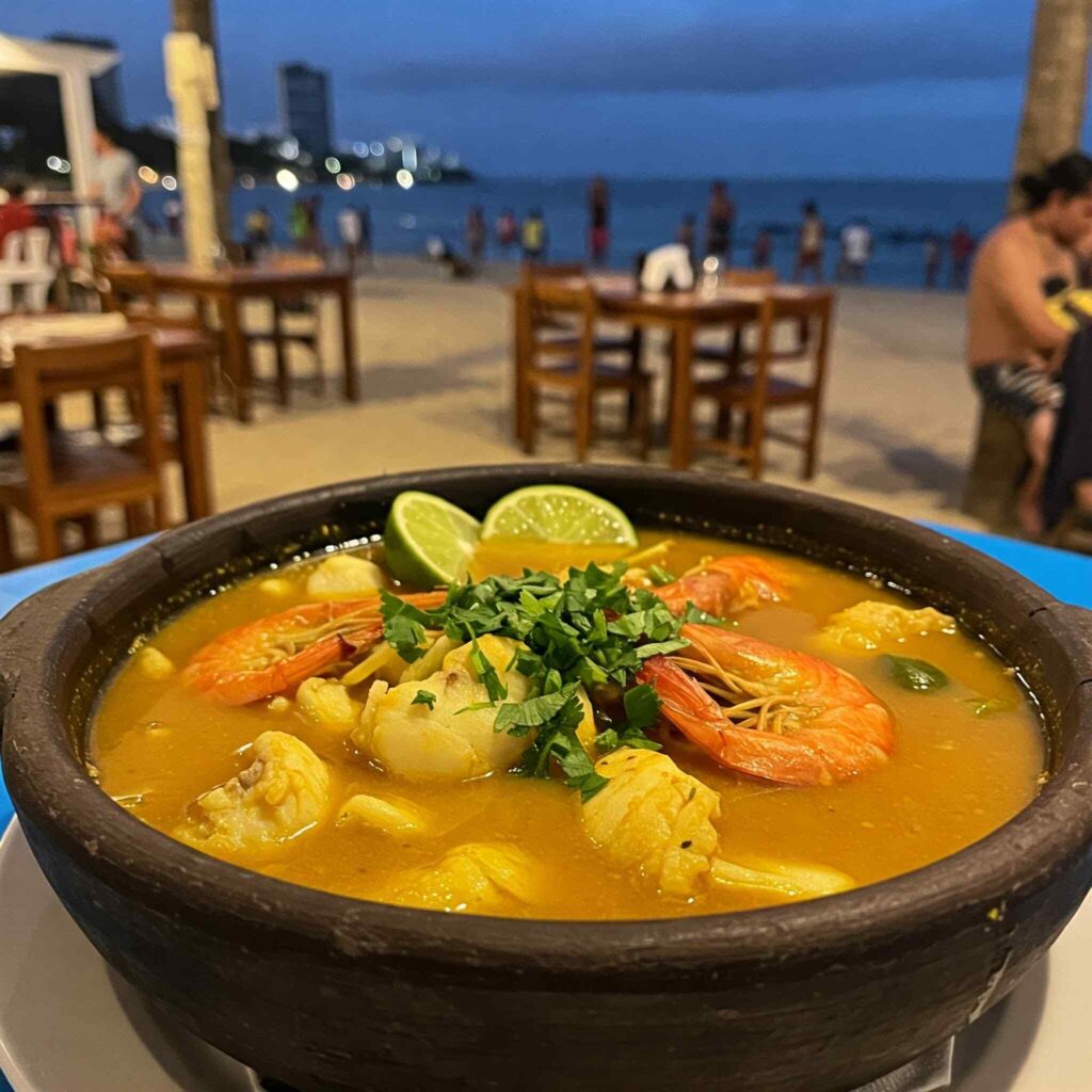 Close-up of a steaming bowl of moqueca, a Brazilian seafood stew, garnished with shrimp, white fish, lime wedges, and cilantro, served at an outdoor beachside restaurant at dusk.