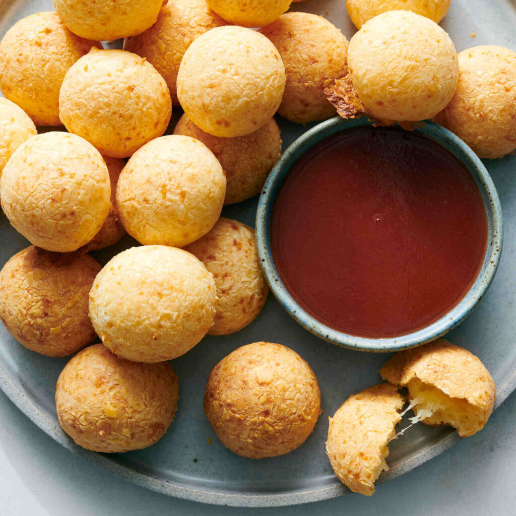 Close-up of pão de queijo (Brazilian cheese bread) on a plate with a bowl of red sauce.
