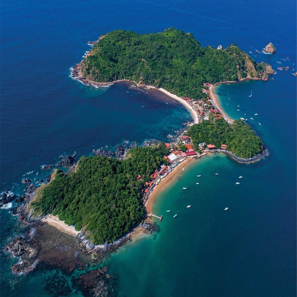 Aerial view of a lush, green island connected by a narrow strip of land with beaches and small buildings, surrounded by turquoise water.
