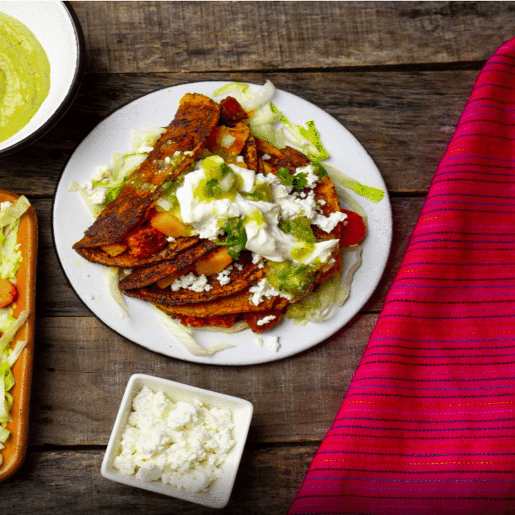 Plate of enchiladas potosinas topped with cream, cheese, and salsa verde on a wooden table with accompaniments.