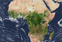 Satellite view of the continent of Africa centered in the frame, surrounded by the blue expanse of the Atlantic and Indian Oceans. Clouds swirl in the atmosphere above the water. The Sahara Desert dominates the northern part of the continent, appearing as a vast expanse of light tan. Moving south, vibrant green hues indicate tropical rainforests and savannas around the equator. The Great Rift Valley is visible as a series of dark lines and elongated lakes in East Africa. The southern tip of the continent shows a mix of green and brown vegetation. The Arabian Peninsula is visible to the northeast, separated by the Red Sea.