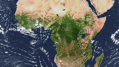 Satellite view of the continent of Africa centered in the frame, surrounded by the blue expanse of the Atlantic and Indian Oceans. Clouds swirl in the atmosphere above the water. The Sahara Desert dominates the northern part of the continent, appearing as a vast expanse of light tan. Moving south, vibrant green hues indicate tropical rainforests and savannas around the equator. The Great Rift Valley is visible as a series of dark lines and elongated lakes in East Africa. The southern tip of the continent shows a mix of green and brown vegetation. The Arabian Peninsula is visible to the northeast, separated by the Red Sea.