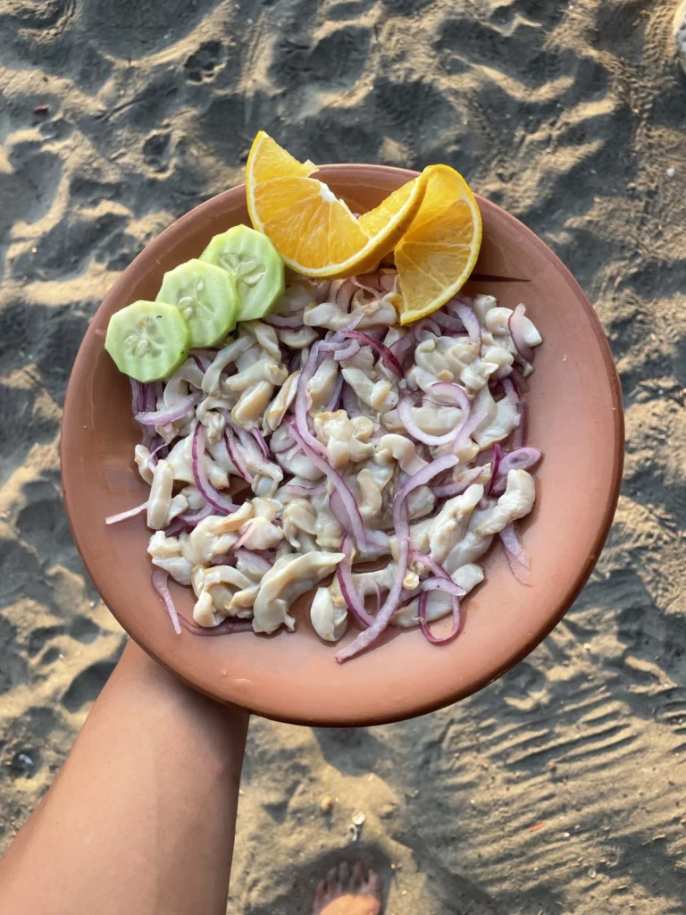plate of tiritas de pescado with orange slices and cucumber on a sandy surface, held by a hand.