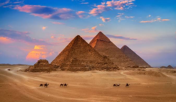 A scenic view of the Great Pyramids of Giza in Egypt, with camels and people in the foreground.